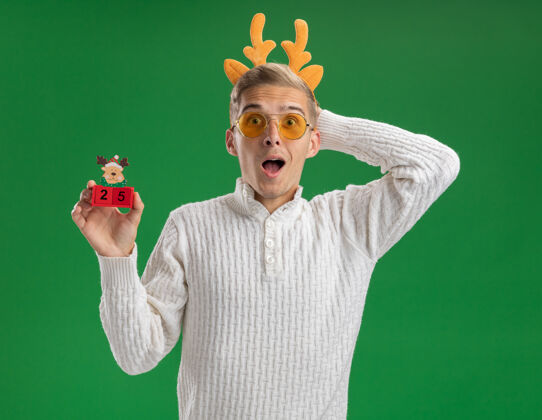 英俊令人印象深刻的年轻帅哥戴着驯鹿鹿角头带戴着眼镜拿着圣诞树玩具和日期保持手后面的头隔离在绿色的墙上年轻驯鹿印象深刻