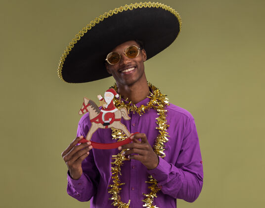 花环微笑着的年轻的非洲裔美国人戴着墨西哥帽子 戴着眼镜 脖子上戴着金箔花环 抱着摇摇木马上的圣诞老人 看着橄榄绿背景上孤立的摄像机墨西哥人眼镜帽子