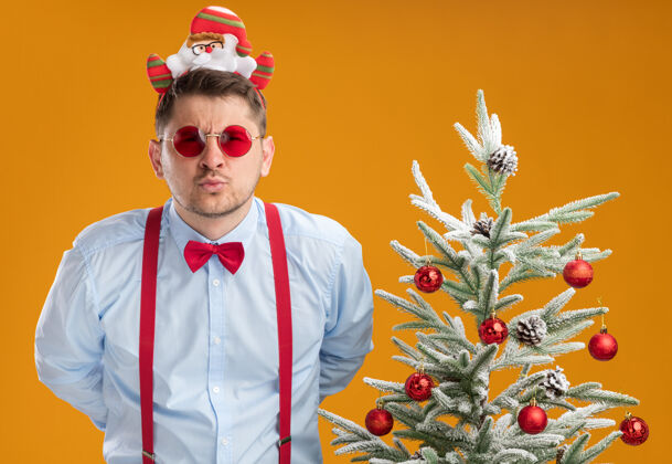 圣诞节戴吊带领结的年轻人站在圣诞树旁 戴着圣诞老人和红眼镜 在橙色背景下严肃地看着摄像机圣诞树轮辋圣诞老人