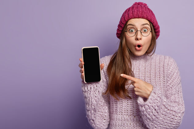 手机表情惊艳的美女 指着手机的空屏 展示自由空间 展示现代电子设备 穿着冬装 隔离在紫色的墙壁上帽子眼镜室内