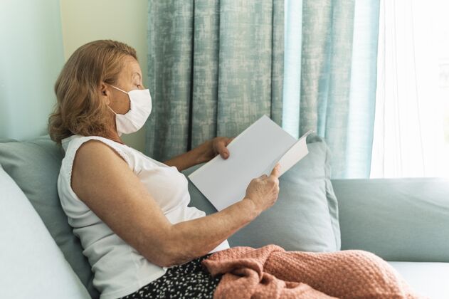 社会距离大流行期间戴着医用口罩的老太太在家看书的侧视图冠状病毒老年人预防