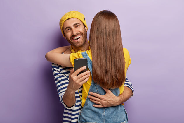 手机快乐的男性年轻人在网上聊天的时间与少女 拥抱妇女谁站在背后的镜头 享受生活 检查信息的追随者媒体在一起手机