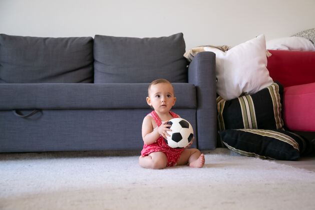 孩子体贴的女婴手里拿着足球 赤脚坐在地毯上 望着别处穿着红色工装裤的可爱的蹒跚学步的孩子在沙发旁的家里玩耍假日 周末和童年的概念可爱工作服小