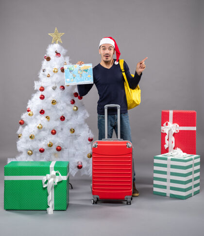 礼物带着黄色背包的兴奋的年轻人拿着地图在灰色的白色圣诞树旁购物年轻背包