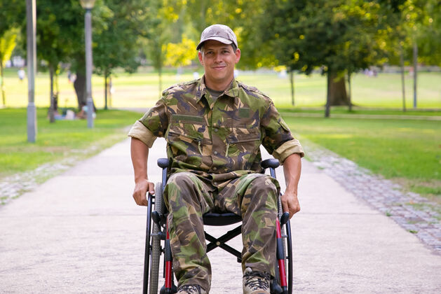 移动快乐的残疾军人在轮椅上穿着迷彩服 走在城市公园的人行道上正面视图老兵的战争或残疾概念高加索受伤成人