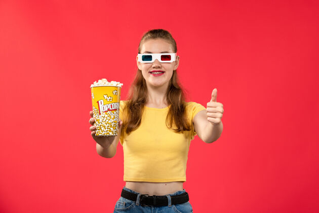 观影的前视图年轻的女性在电影院拿着爆米花戴着d太阳镜在红墙电影院看电影的女性乐趣时间电影漂亮的电影院成人的