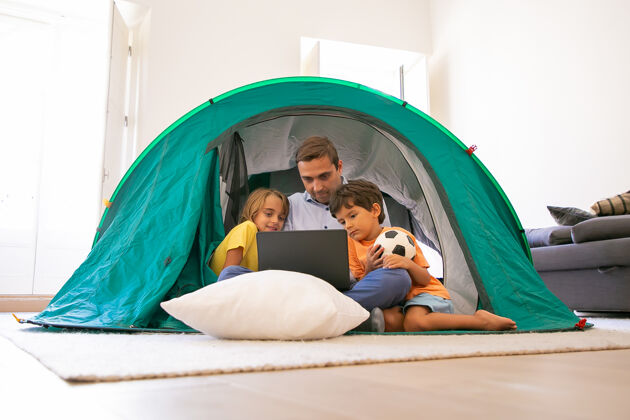 女儿有爱心的爸爸和孩子们盘腿坐在家里的帐篷里 看着笔记本电脑屏幕可爱的孩子们和爸爸一起在电脑上看电影童年 家庭时间和周末的概念男孩关心盘腿