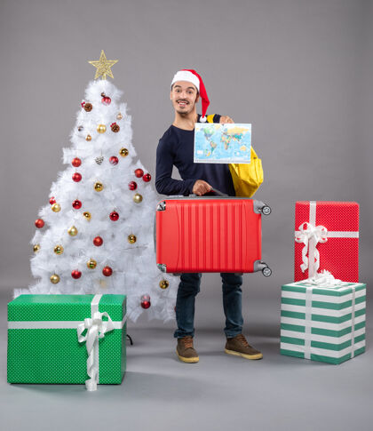 快乐快乐的年轻人背着背包 拿着圣诞树旁的地图和灰色的礼物圣诞老人圣诞树快乐的年轻人