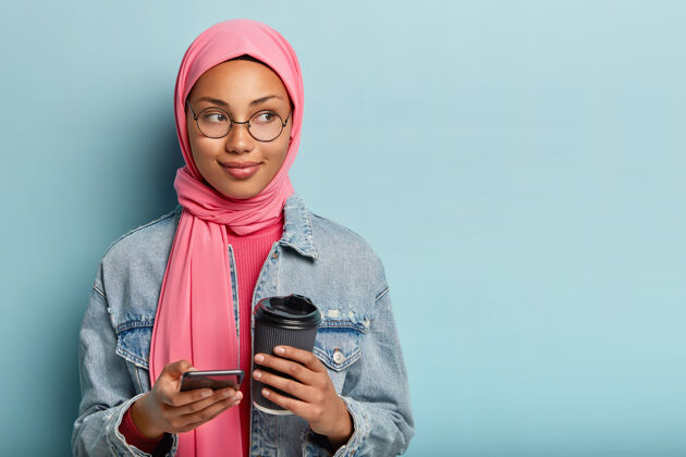 短信科技和通讯理念身着粉色面纱的妇女的照片 使用新安装的智能手机应用程序 端着咖啡走 戴着圆眼镜 站在室内蓝色墙壁上宗教个性业余时间