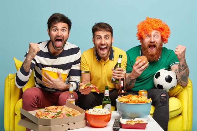 足球快乐的三个朋友成为足球迷 看足球 在客厅的沙发上摆姿势 吃快餐 喝冷啤酒酒精比赛享受