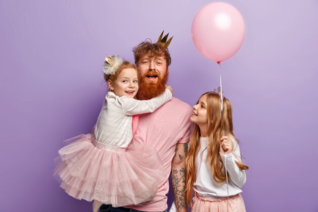 孩子家庭 父母和节日的概念有趣的父亲招待孩子 手上抱着小女儿 在聚会前装饰房间 穿喜庆的衣服 隔离在紫色的墙壁上氦红发未刮胡子