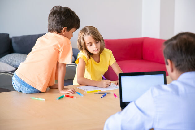 成人白种人爸爸在笔记本电脑上工作 可爱的孩子在桌上画涂鸦专注的金发女孩和马克一起画画 哥哥看着她童年 创造力和周末的概念朋友男人可爱