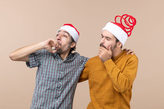 视图前视图两个戴着圣诞帽的体贴的家伙在米色孤立的背景下看着什么东西微笑男人伙计们
