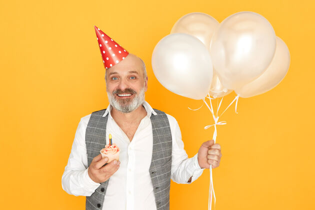 乐趣快乐快乐的老年男性肖像 留着胡须 穿着优雅的衣服 戴着圆锥形帽子 举着生日蛋糕和氦气球帽子聚会姿势