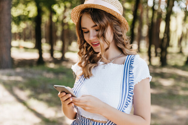 移动迷人的卷发女孩穿着时髦的衣服看着手机屏幕户外拍摄迷人的女模特戴着帽子在公园拍照后发短信户外小工具智能手机