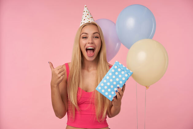 迷人快乐的年轻金发女郎 随意的发型 粉色上衣 生日圆锥形帽 站在粉色背景下 一堆五彩的氦气球 看着相机 高兴地竖起大拇指无包装上衣微笑