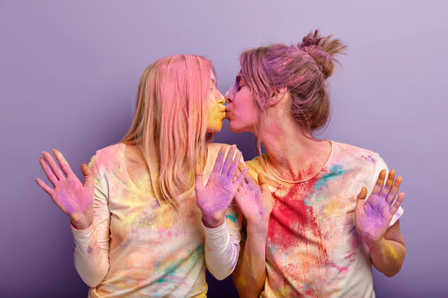 情感年轻的好朋友女性深情地亲吻 一起玩乐 涂上彩粉 庆祝胡里节 展示彩绘的手掌或明亮的手臂 对着紫色的墙壁摆姿势室内女同性恋充满活力