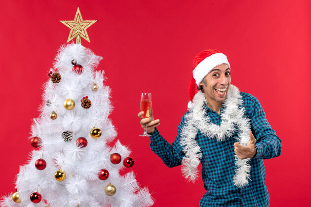 新年前夜圣诞心情与微笑的年轻人圣诞老人帽子在一个蓝色的衬衫剥离举行一杯葡萄酒附近圣诞树庆祝圣诞帽人