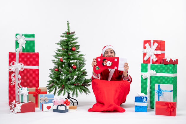 礼物微笑的圣诞老人坐在地上 把圣诞袜举到他脸上 礼物和装饰在白色背景上的新年树旁圣诞老人圣诞盒子