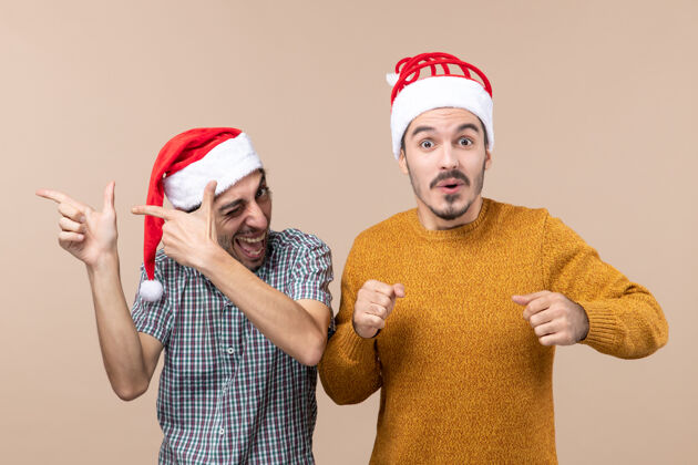 圣诞老人前视图两个戴着圣诞帽的快乐的家伙一个眨着眼睛 另一个在米色孤立的背景上用窥视的眼睛米色眨眼两个快乐的家伙