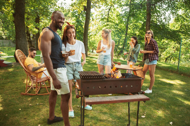 烧烤一群快乐的朋友在阳光明媚的日子里举行啤酒和烧烤派对一起在户外的森林空地或后院休息欢庆和放松 欢笑夏日生活方式 友谊理念午餐自然一起