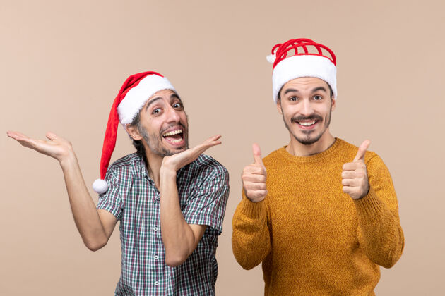 帽子正面图两个带着圣诞帽的微笑的家伙 一个在展示什么 另一个在米色背景上用双手做着竖起大拇指的手势前面男人两个