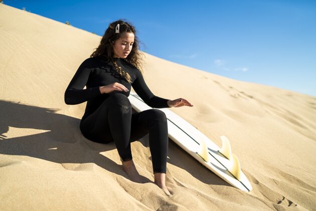 休闲浅聚焦拍摄一个迷人的女性坐在一个沙山与冲浪板在一边活跃姿势度假村