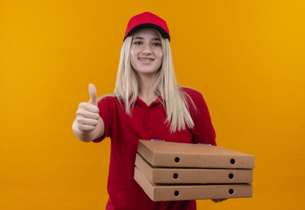 送货微笑的小女孩穿着红色t恤 戴着帽子 戴着牙套 拿着披萨 在孤立的橙色背景上竖起大拇指微笑红色帽子