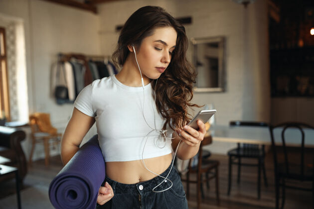 曲线穿着牛仔裤和紧身上衣的运动型棕褐色加大码年轻女性 腋下夹着卷起的健身垫 在手机上输入短信训练放松能量
