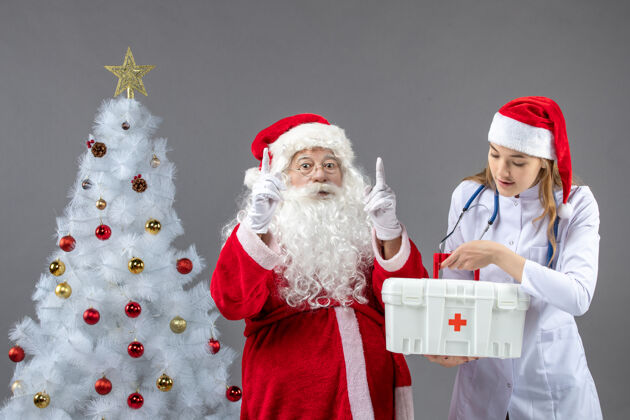 人们圣诞老人和女医生的前视图 女医生拿着急救箱在灰色的墙上十二月健康举行