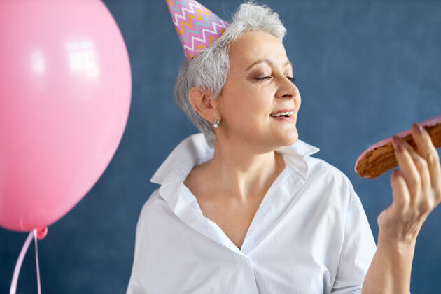 Eclair在生日派对上 身着时尚白衬衫 戴着圆锥形帽子 手持粉色氦气球的快乐退休女士随着音乐翩翩起舞老年人成熟吃