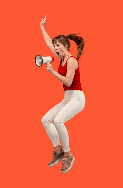 喊叫美丽的年轻女子用扩音器在红色背景上孤立地跳跃奔跑的女孩在运动或运动人类的情感和面部表情的概念表情沟通跳跃