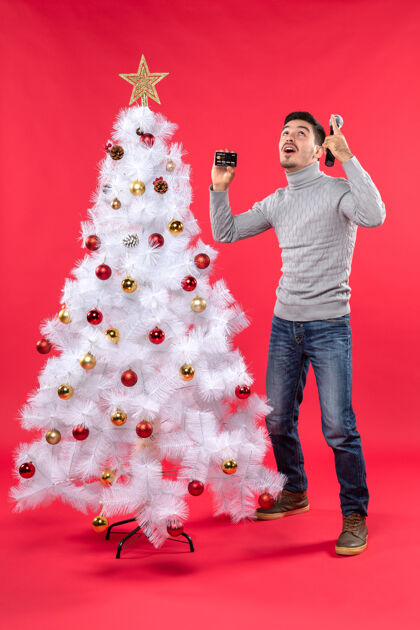 圣诞圣诞气氛 年轻人站在装饰好的圣诞树旁 拿着麦克风和电话往上看礼物微笑电话