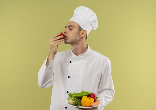 盘子站在剖面图年轻的男厨师穿着厨师制服拿着盘子上的蔬菜和闻番茄男蔬菜西红柿