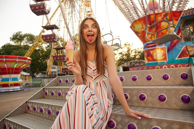 公园有趣可爱的年轻女子穿着夏装在游乐场摩天轮上摆姿势 手里拿着冰激凌 露出粉红色的舌头衣服美丽长