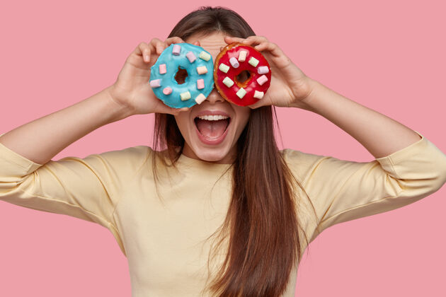美味的快乐的年轻女子拿着两个美味的甜甜圈抵着眼睛 惊讶地张开嘴 享受甜点 隔离在粉红色的空间里零食黑色头发吃