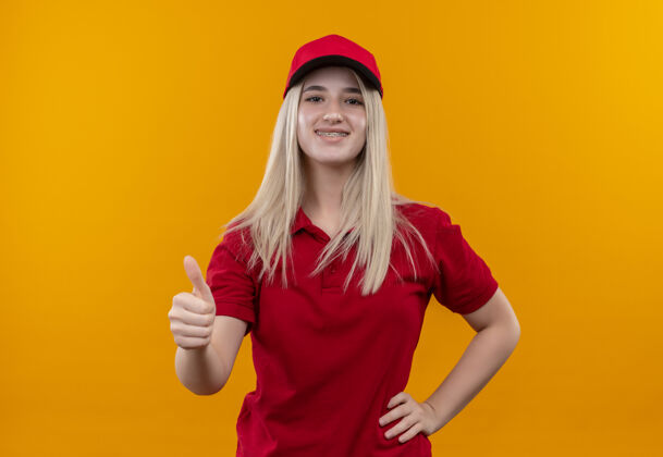 红色带着微笑的小女孩穿着红色t恤 戴着戴着牙套的帽子 手放在臀部 拇指朝上放在孤立的橙色背景上帽子手臀部