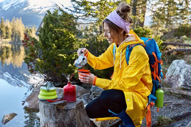 制作心满意足的女游客从茶杯里的咖啡机倒咖啡 用红色的丁烷瓶 用背包穿雨衣茶浇注自然
