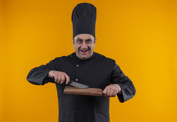 刀快乐的中年男厨师 身着厨师制服 手持黄色墙上刻有菜板的小刀制服快乐中年