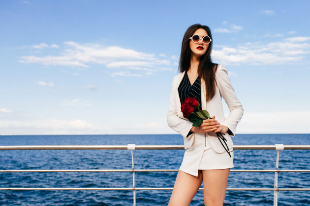 地平线年轻的时髦的黑发女子在堤岸摆姿势与小红色的玫瑰花束苍蝇美丽热