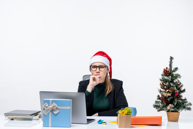 圣诞老人一位体贴的年轻女士 戴着圣诞老人的帽子 坐在一张桌子旁 桌子上放着圣诞树和礼物 在白色背景上指着上方笔记本电脑年轻女人
