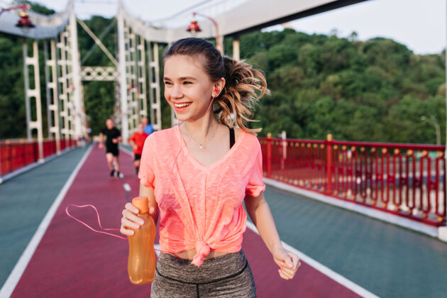夏天笑容可掬的金发女孩拿着一瓶果汁 沿着煤渣跑道奔跑神奇的女模特在体育场玩得很开心人微笑苗条