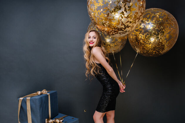 时尚快乐的聚会时间令人惊奇的快乐的年轻女子穿着黑色的豪华礼服 长卷曲的金发拿着装满金色金属箔的大气球礼物 微笑 心情愉快迷人女人圣诞节