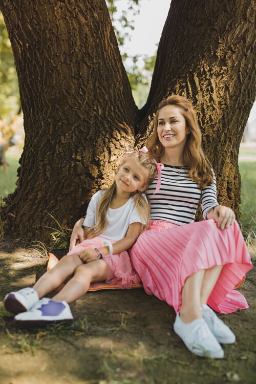 收养粉红色的裙子微笑的爱的女人穿着粉红色的裙子坐在她时髦的小女儿旁边一起坐着树