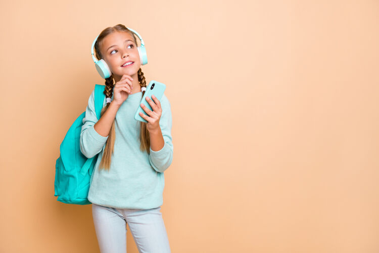 套头衫美丽的小女孩的照片听耳机最喜欢的歌走路学校举行电话想周末的梦想穿蓝色套头衫牛仔裤隔离米色墙情人耳机时尚