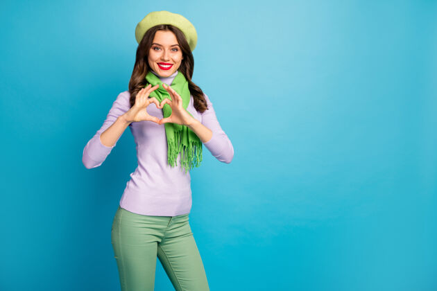 天照片中搞笑迷人的女士手牵手心形象征心意穿着时尚的绿色贝雷帽紫色高领围巾长裤孤立的蓝色墙壁情人喜欢休闲