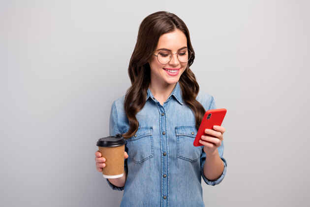 阅读美丽的自由职业者的照片女士拿着纸杯热咖啡饮料工作休息浏览电话阅读后穿规格休闲牛仔裤牛仔衬衫孤立的灰色空闲时间互联网商务女性