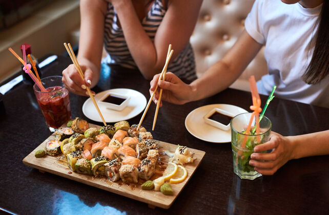 黄瓜两个杨家的女孩在餐厅吃着美味的卷寿司 用筷子 酱油和鸡尾酒装在木板上盘子芥末饮食