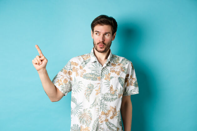 年轻度假时穿着夏装的帅哥 指着空荡荡的空间 做着惊异的脸 站在蓝色的背景上指着衬衫男性