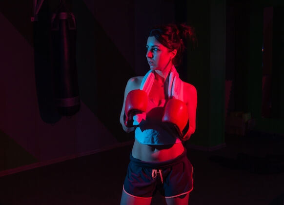 运动年轻女子拳击手戴着拳击手套 肩上扛着毛巾 在黑暗的墙上霓虹灯渐变着红蓝色的灯光防守运动霓虹灯